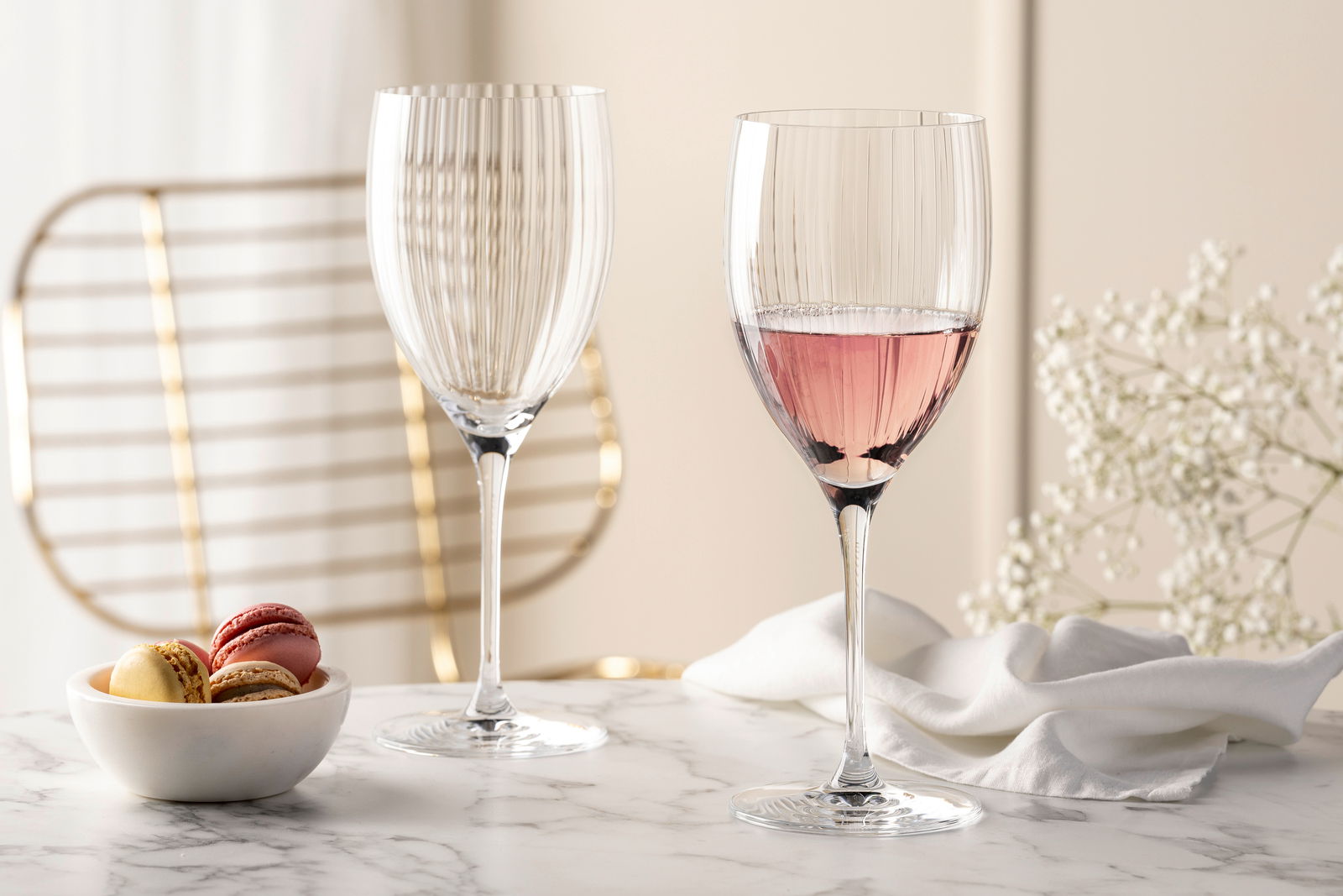 Zwei elegante Rotweingläser POESIA 600ml auf einem Marmortisch, eines gefüllt mit roséfarbenem Wein, im Hintergrund eine goldene Stuhllehne und weiße Blumen, seitliche Perspektive.