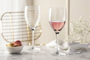 Zwei elegante Rotweingläser POESIA 600ml auf einem Marmortisch, eines gefüllt mit roséfarbenem Wein, im Hintergrund eine goldene Stuhllehne und weiße Blumen, seitliche Perspektive.