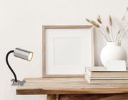 Klemmstrahler aus Metall in Nickeloptik, seitliche Perspektive, befestigt an einem Holztisch, neben einem Bilderrahmen und einer Vase.