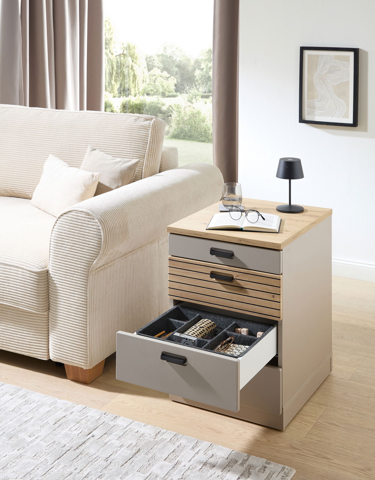 Rollcontainer mit geöffneten Schubladen, gefüllt mit Büroartikeln, neben einem Sofa in einem Wohnzimmer. Perspektive von vorne.