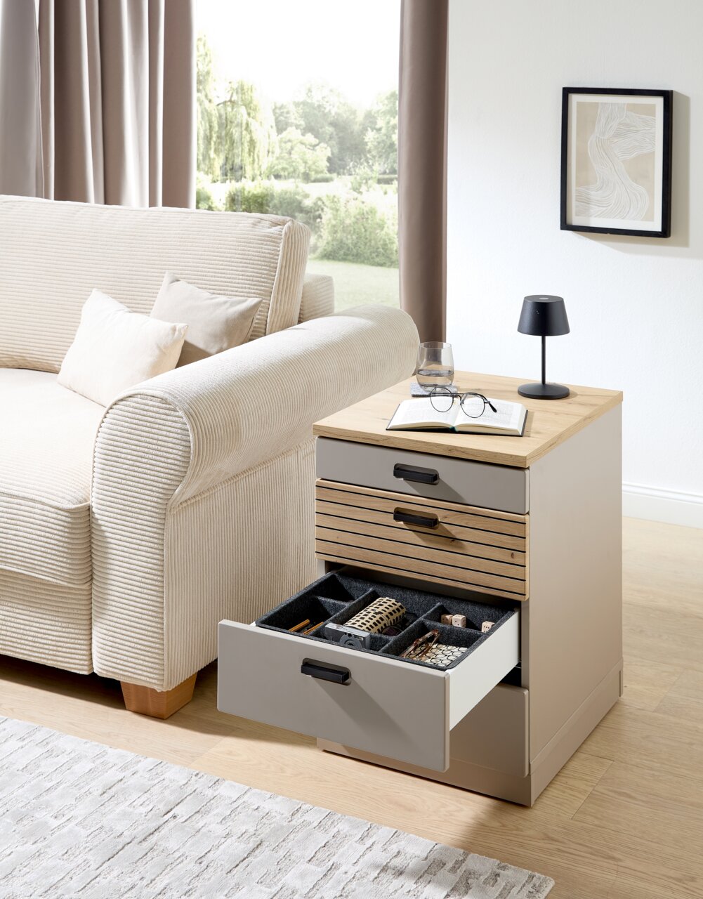 Rollcontainer mit geöffneten Schubladen, gefüllt mit Büroartikeln, neben einem Sofa in einem Wohnzimmer. Perspektive von vorne.