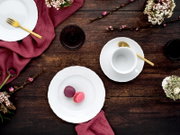 Draufsicht auf ein elegantes Tischgedeck mit weißem Porzellan (Teller und Tasse), goldfarbenem Besteck, zwei Macarons und bordeauxrotem Stoff auf rustikalem Holztisch mit Blumen