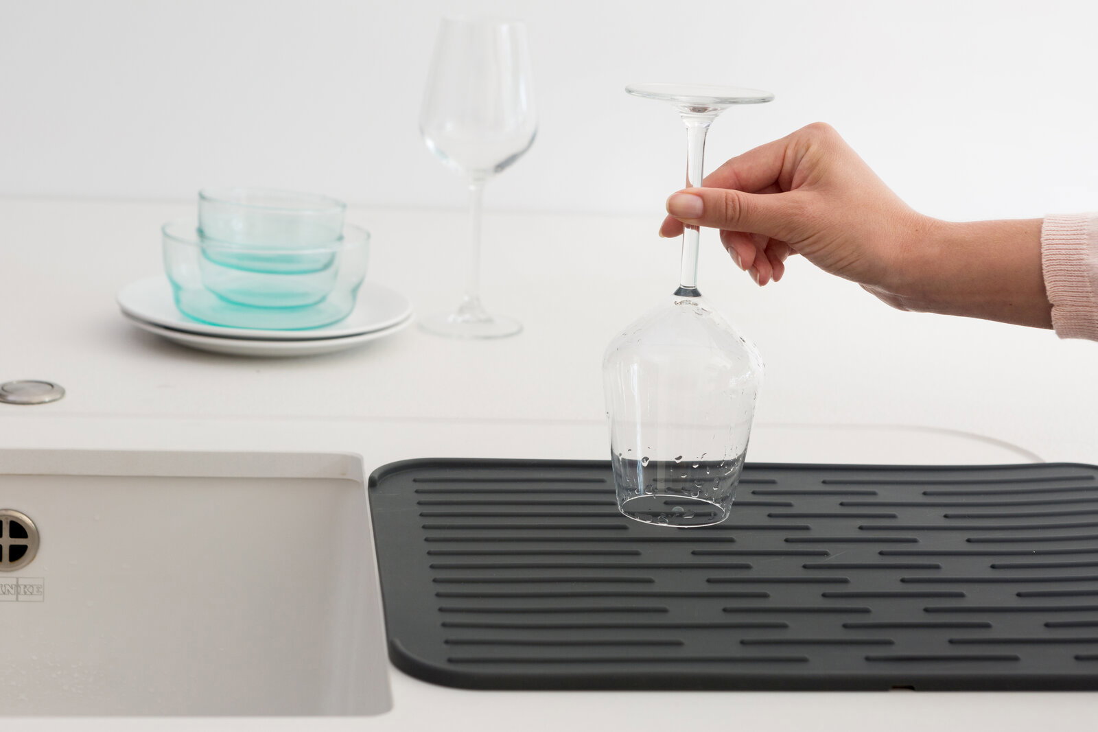 Brabantia Abtropfmatte SINKSIDE Eine schwarze Abtropfmatte neben einem Waschbecken, auf der ein Weinglas von einer Hand gehalten wird. Im Hintergrund sind weitere Gläser und Schalen zu sehen. Perspektive von oben.