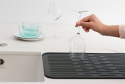 Eine schwarze Abtropfmatte neben einem Waschbecken, auf der ein Weinglas von einer Hand gehalten wird. Im Hintergrund sind weitere Gläser und Schalen zu sehen. Perspektive von oben.