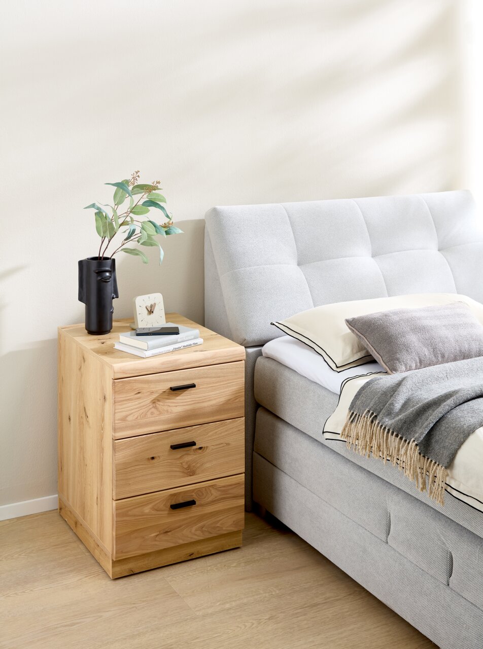 Holz-Rollcontainer mit drei Schubladen, neben einem Bett platziert, aus seitlicher Perspektive fotografiert.