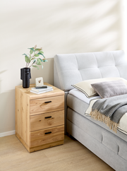 Holz-Rollcontainer mit drei Schubladen, neben einem Bett platziert, aus seitlicher Perspektive fotografiert.