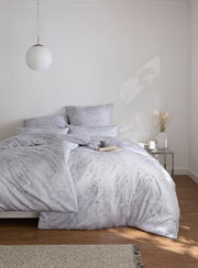 Bettwäsche GREGORIO in einem hellen Schlafzimmer, frontal fotografiert, mit elegantem Muster und dekorativen Kissen.