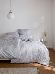 GREGORIO-beddengoed in een lichte slaapkamer, gefotografeerd van voren, met een elegant patroon en decoratieve kussens.