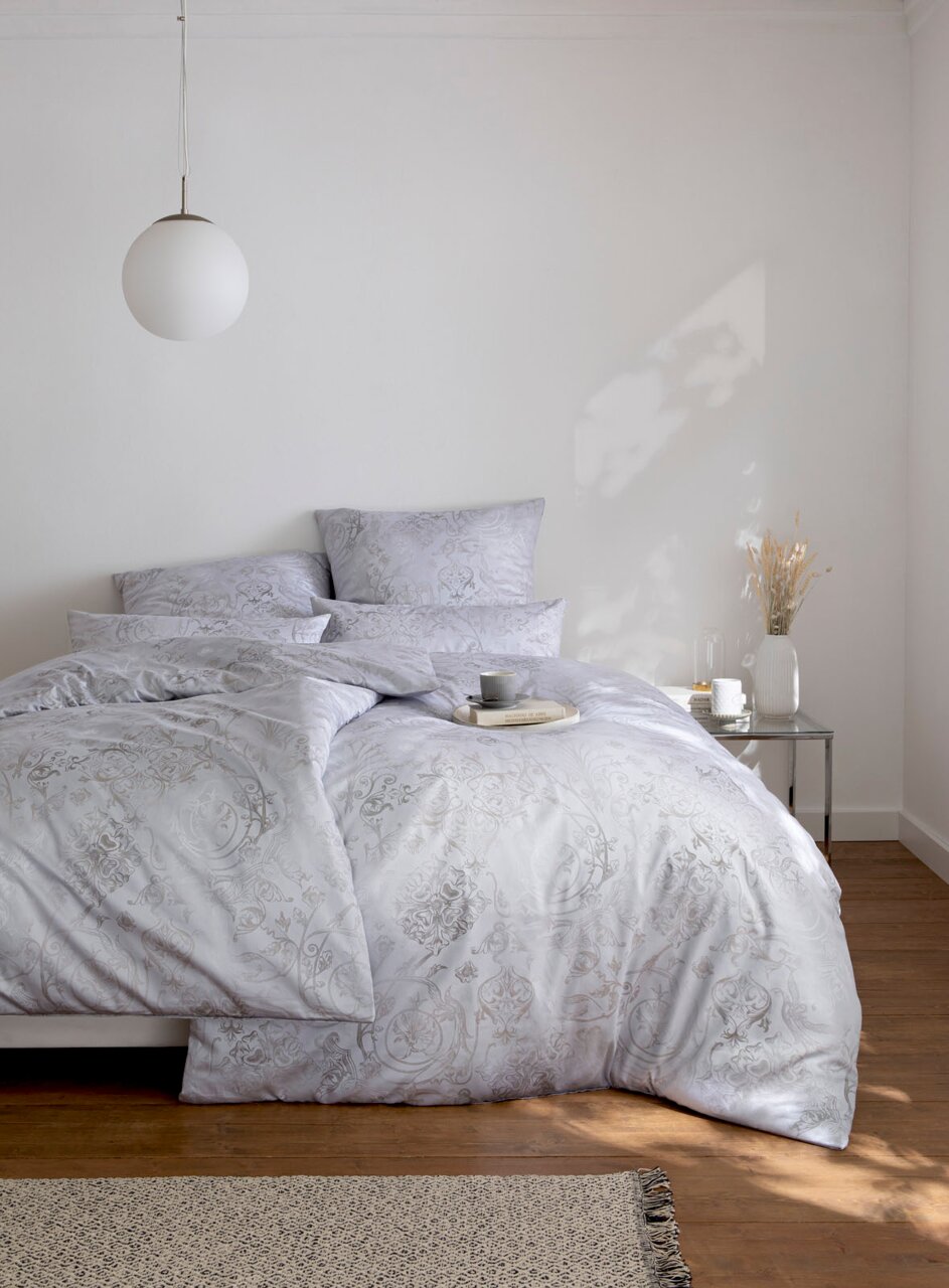GREGORIO-beddengoed in een lichte slaapkamer, gefotografeerd van voren, met een elegant patroon en decoratieve kussens.