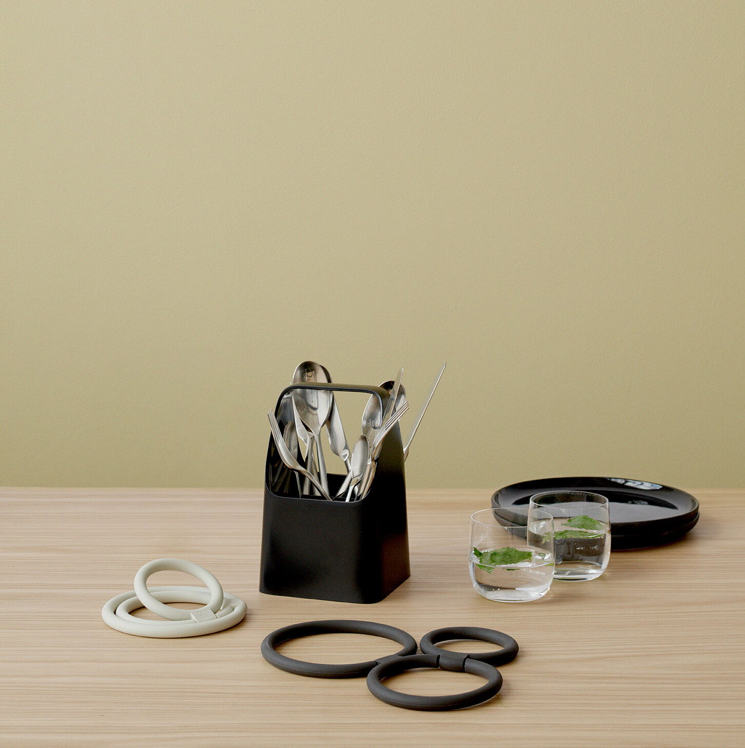 Schwarze Aufbewahrungsbox mit Besteck, von vorne gesehen, auf einem Holztisch mit weiteren Küchenutensilien.