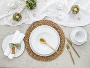 Draufsicht auf eine stilvolle Tischdekoration mit weißem Geschirr auf geflochtenem Platzset, goldfarbenem Besteck, Leinenläufer, Kerzen und kleinen Herzdetails