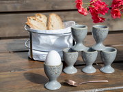 Graue, gesprenkelte Eierbecher aus Keramik (Set) auf rustikalem Holztisch, ein Eierbecher mit Ei, daneben Brotkorb und Löffel; Perspektive: leicht schräg von vorne auf Tischhöhe