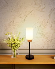 Moderne Tischlampe mit zylindrischem, milchig-weißem Glasschirm, Holzdetail und schwarzem Standfuß auf Holzkonsole neben Glasvase mit grünem Blumenstrauß vor strukturierter Wand, frontal aufgenommen