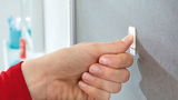 Nahaufnahme einer Hand, die einen tesa Powerstrip Waterproof an einer grauen Wand befestigt.