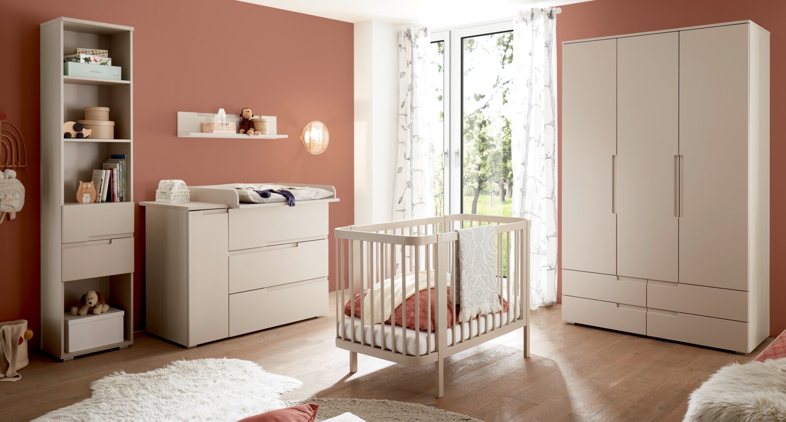 Helles Babyzimmer mit weißem Babybett, Wickelkommode, Regal und großem Kleiderschrank vor terrakottafarbener Wand, aufgenommen in frontaler Perspektive mit leichtem Weitwinkel