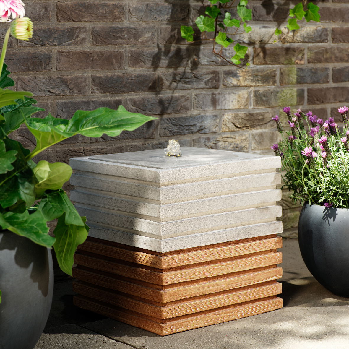 Lumineo Brunnen eckig Ribbel Outdoor Eckiger Brunnen aus gestapelten, gerippten Platten in einem Garten, umgeben von Pflanzen, seitliche Perspektive.