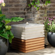 Eckiger Brunnen aus gestapelten, gerippten Platten in einem Garten, umgeben von Pflanzen, seitliche Perspektive.