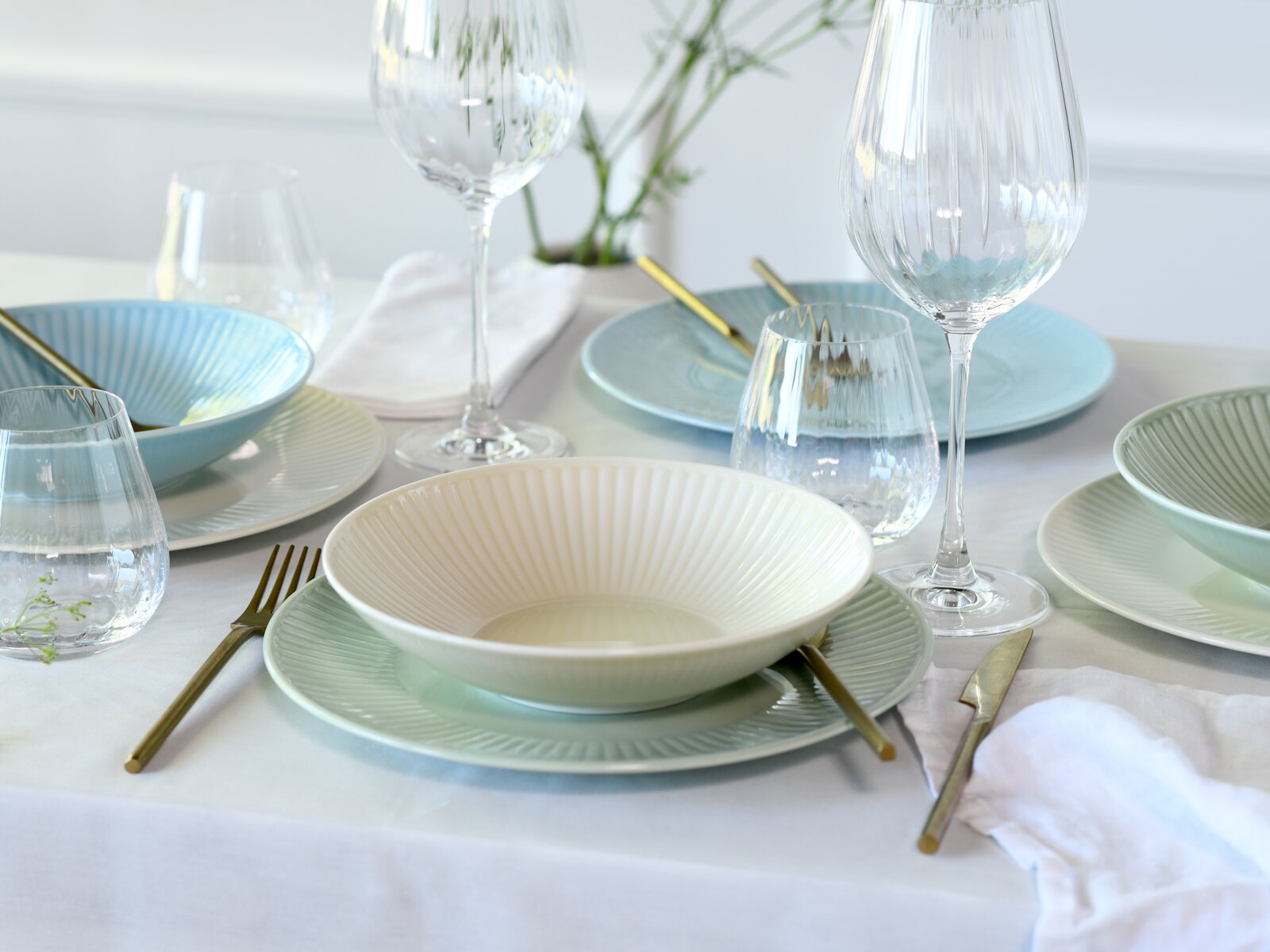 Schlicht gedeckter Esstisch mit geriffeltem Geschirr in Pastellfarben (Schale in Creme auf grünem Teller, blaue Schalen), goldfarbenem Besteck und klaren Weingläsern, aus leicht schräger Seitenansicht auf Tischhöhe