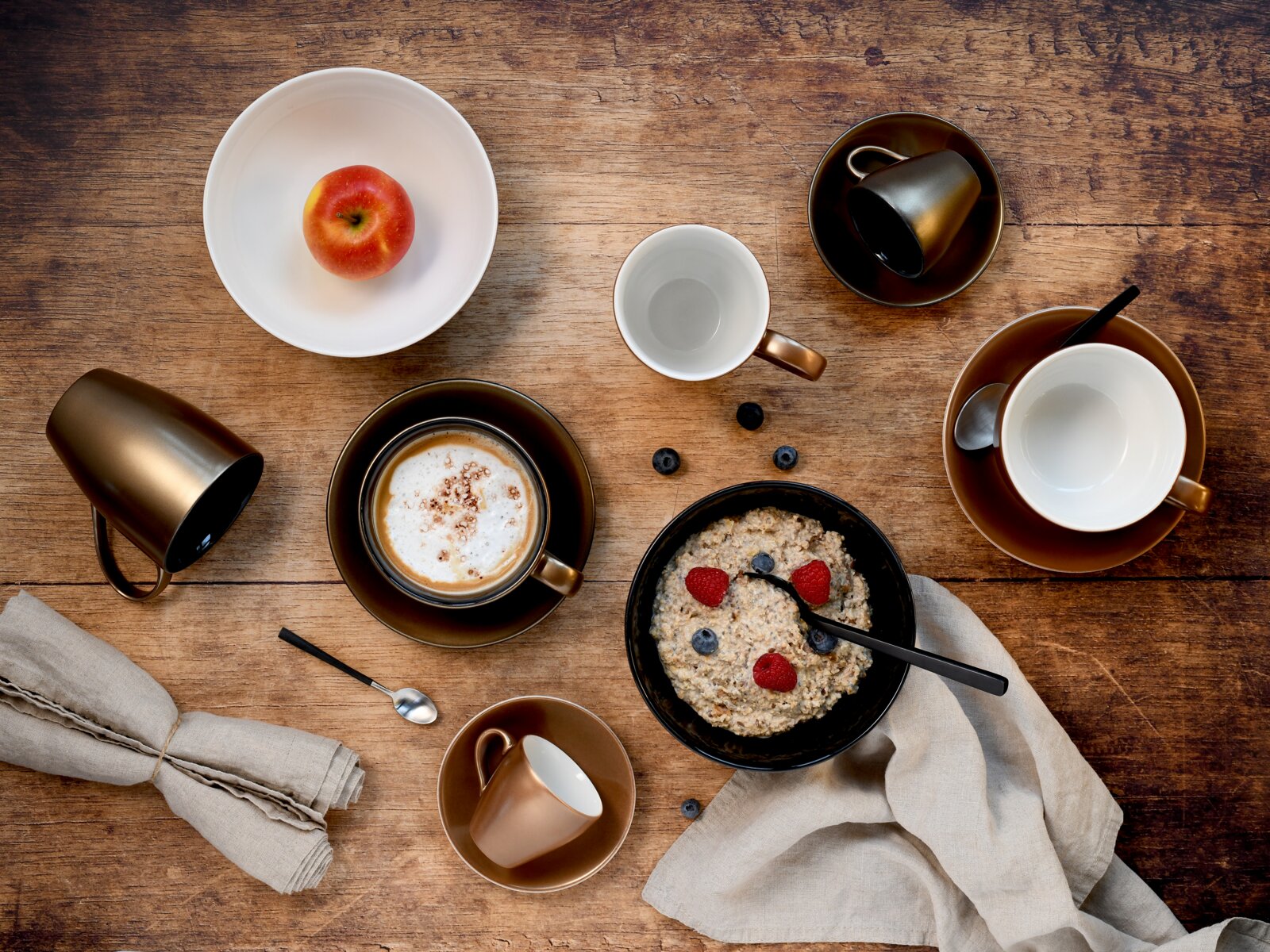Draufsicht auf rustikalen Frühstückstisch aus Holz mit Cappuccino in Tasse, Schale Porridge mit Beeren und Löffel, Apfel in weißer Schüssel, mehreren Tassen und Leinenservietten.