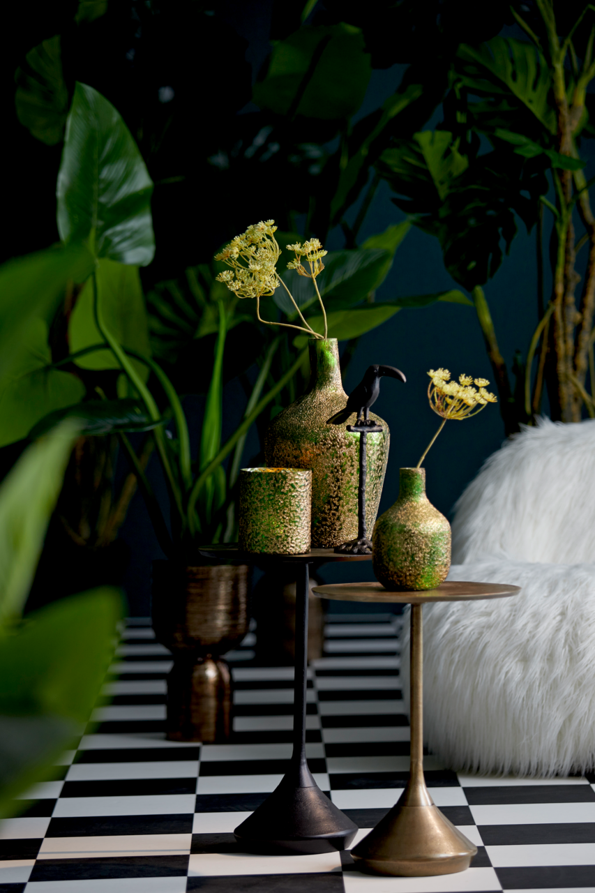Zwei Beistelltische in einem stilvollen Interieur, dekoriert mit grünen und goldenen Vasen und einem schwarzen Vogel, umgeben von üppigen Pflanzen. Perspektive: seitlich.