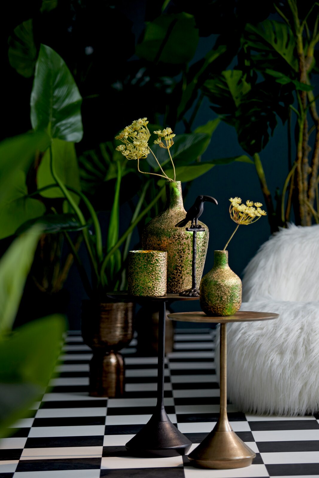 Zwei Beistelltische in einem stilvollen Interieur, dekoriert mit grünen und goldenen Vasen und einem schwarzen Vogel, umgeben von üppigen Pflanzen. Perspektive: seitlich.