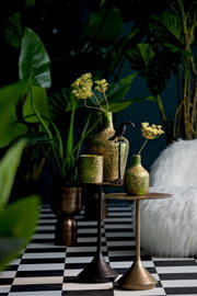 Zwei Beistelltische in einem stilvollen Interieur, dekoriert mit grünen und goldenen Vasen und einem schwarzen Vogel, umgeben von üppigen Pflanzen. Perspektive: seitlich.
