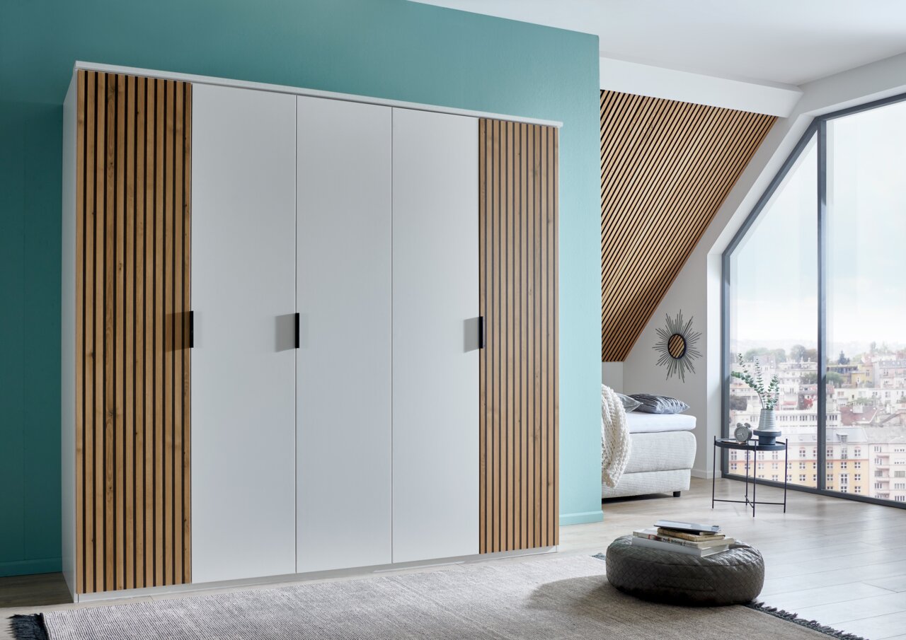 Moderner 5-türiger Drehtürenschrank mit weißen Fronten und Holzlamellen an den Seiten, in einem stilvoll eingerichteten Raum mit Blick auf ein großes Fenster. Perspektive von vorne links.