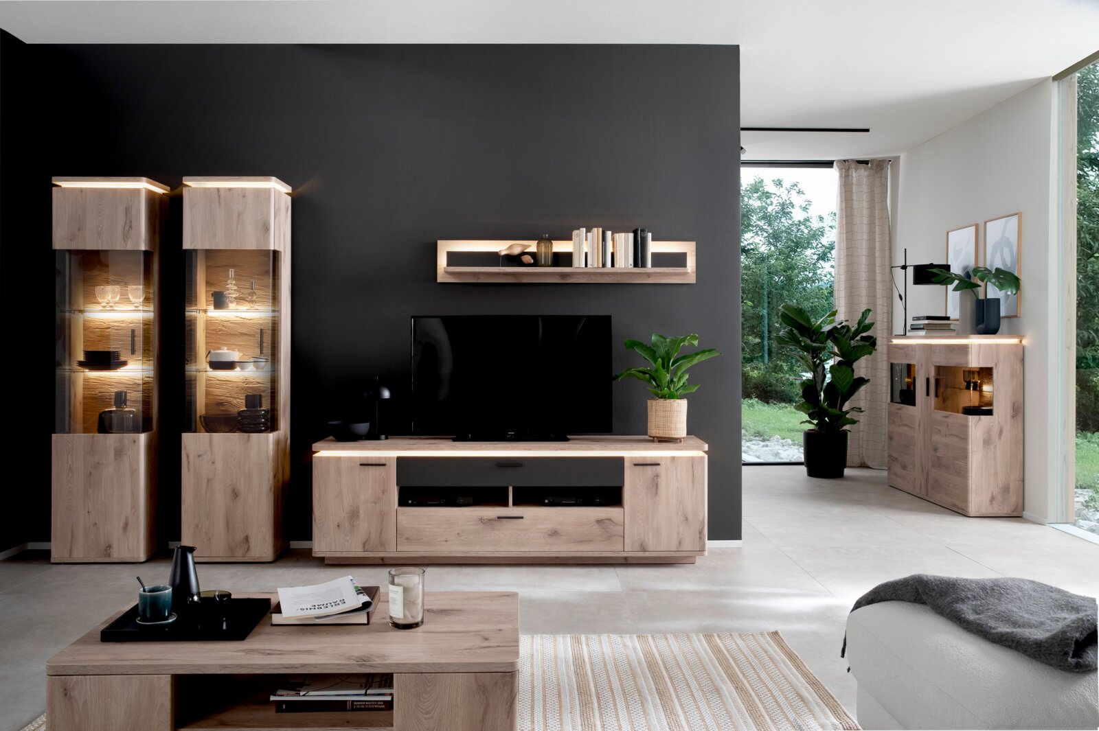Moderne woonkamerset in lichte eikenlook met twee verlichte vitrinekasten, een tv-lowboard, een wandrek en een dressoir tegen een donkere accentmuur; vooraanzicht van de meubelgroep in de kamer.