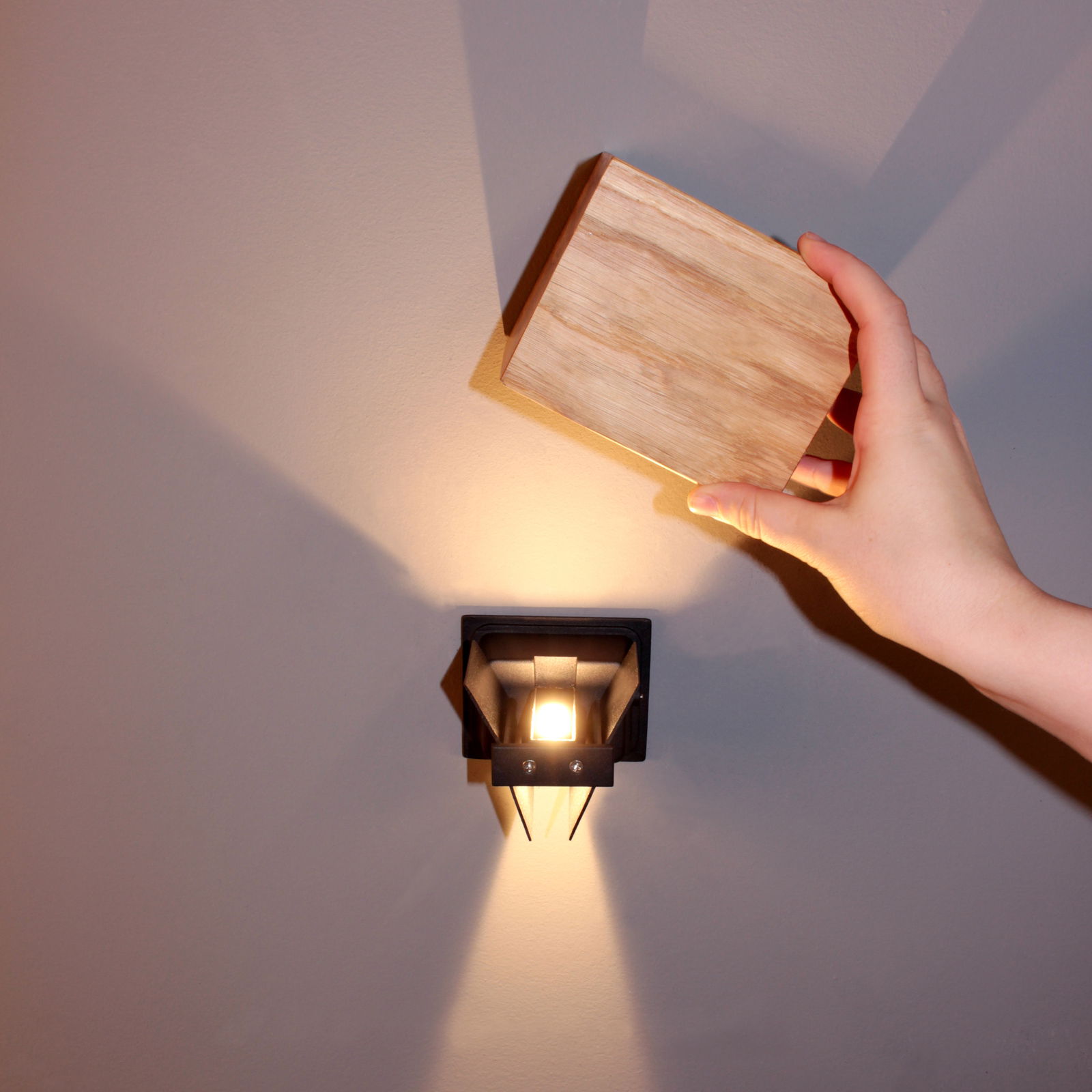 Moderne Wandleuchte mit geometrischem Design, von vorne betrachtet, mit einer Hand, die ein Holzpaneel darüber hält.