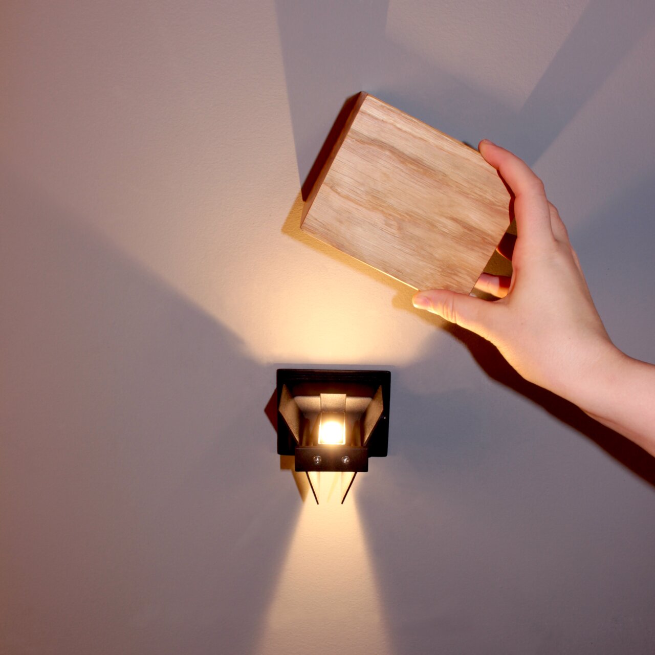 Moderne Wandleuchte mit geometrischem Design, von vorne betrachtet, mit einer Hand, die ein Holzpaneel darüber hält.