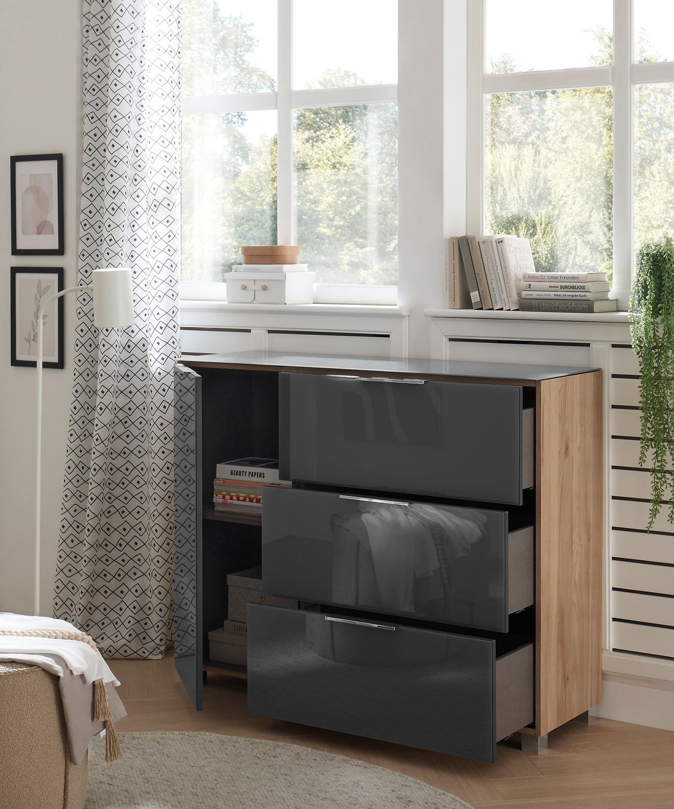 Moderne Kommode mit drei glänzenden, dunklen Schubladen und einer Holztür, seitliche Perspektive, in einem hellen Raum mit großen Fenstern und dekorativen Elementen.