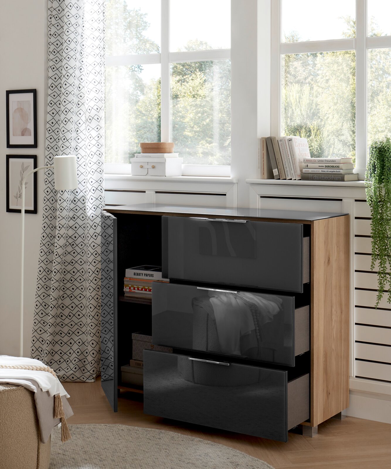 Moderne ladekast met drie glanzende, donkere lades en een houten deur, gezien vanaf de zijkant, in een lichte ruimte met grote ramen en decoratieve elementen.