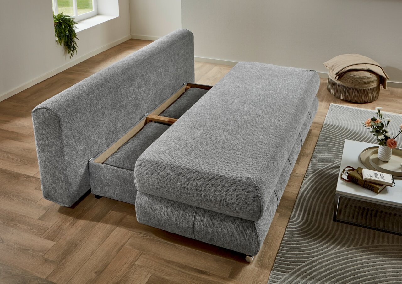 Grijze functionele bank in uitgeklapte stand, zijaanzicht, op een houten vloer met een modern interieur.