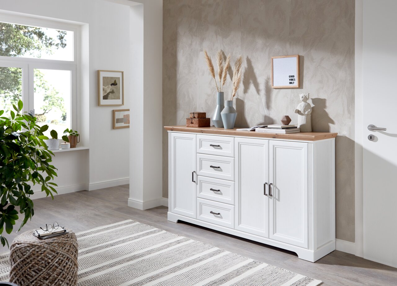 Weißes XL-Sideboard der Serie JUDITH mit Holzplatte, dekoriert mit Vasen und Büchern, in einem modernen Wohnzimmer aus seitlicher Perspektive.
