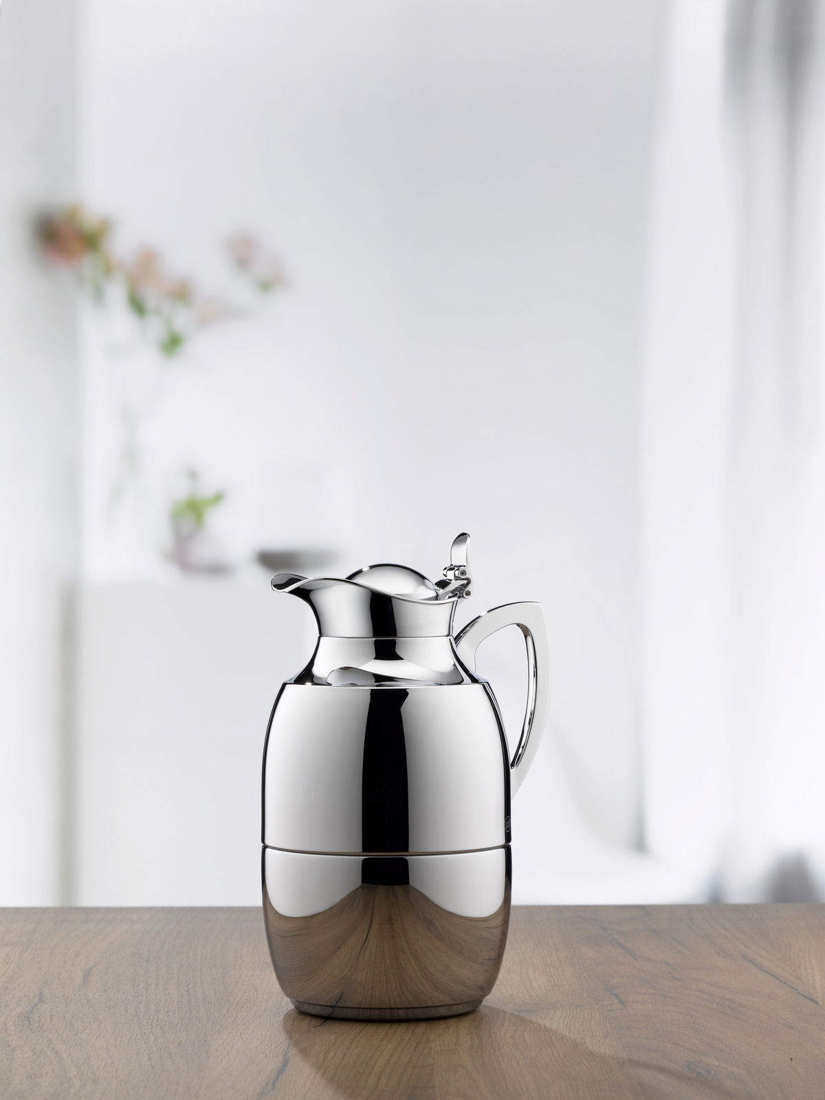 Elegante Isolierkanne Juwel aus Edelstahl, frontal auf einem Holztisch vor einem unscharfen Hintergrund mit Blumen.