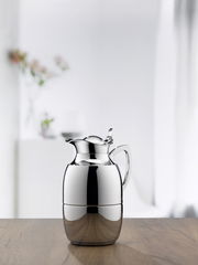 Elegante Isolierkanne Juwel aus Edelstahl, frontal auf einem Holztisch vor einem unscharfen Hintergrund mit Blumen.