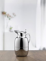 Elegante Isolierkanne Juwel aus Edelstahl, frontal auf einem Holztisch vor einem unscharfen Hintergrund mit Blumen.