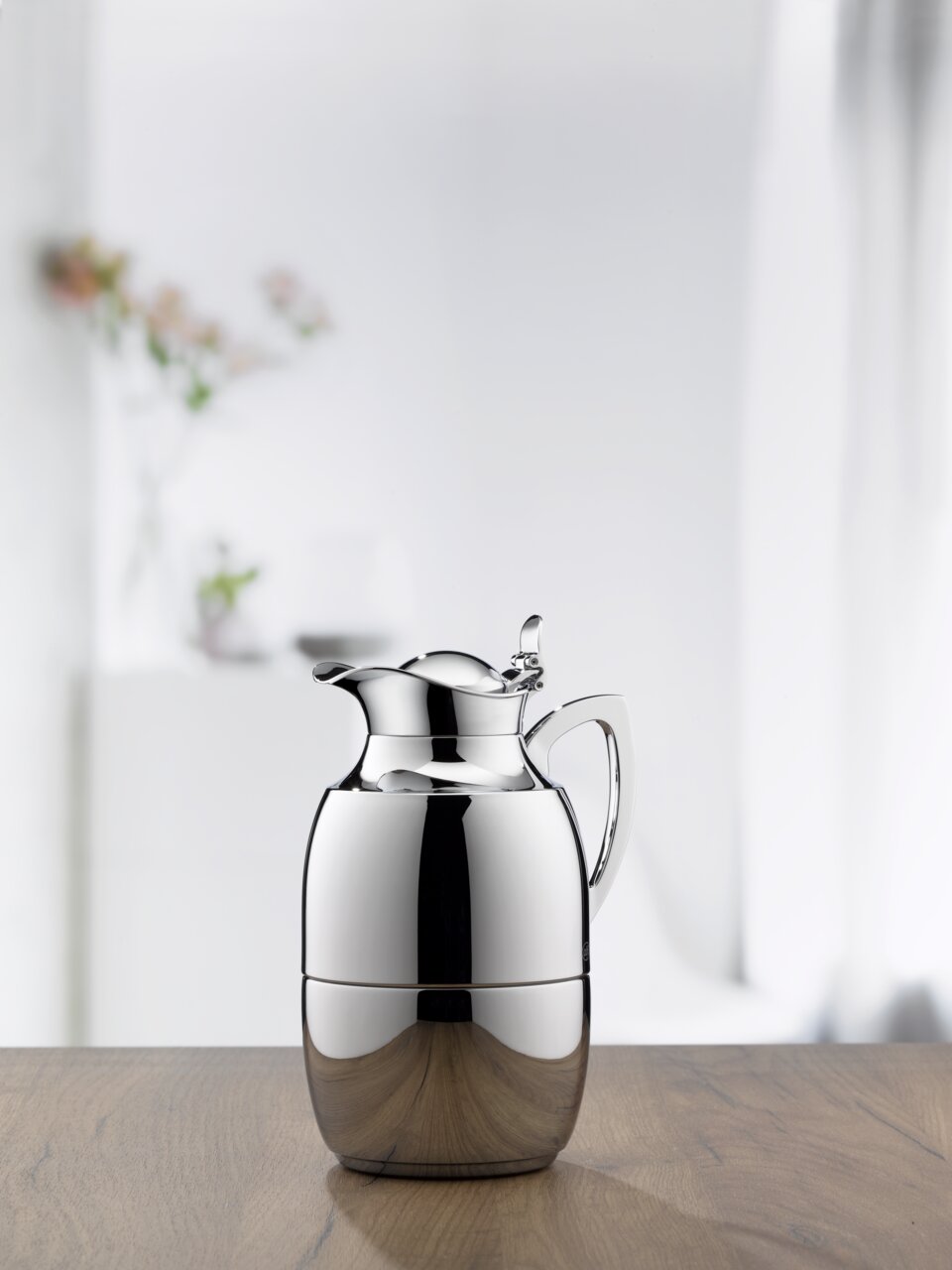 Elegante Isolierkanne Juwel aus Edelstahl, frontal auf einem Holztisch vor einem unscharfen Hintergrund mit Blumen.