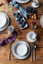 Draufsicht auf einen rustikal gedeckten Holztisch mit grauweißem Keramikgeschirr, Tassen, Besteck, Stoffservietten sowie Schale mit Blaubeeren, Platte mit Pflaumen, Walnüssen und kleinen Blumenvasen.
