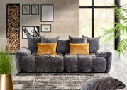 Graues Megasofa mit weichen Kissen und zwei gelben Zierkissen, frontal fotografiert in einem modernen Wohnzimmer mit großen Fenstern und Wanddekorationen.