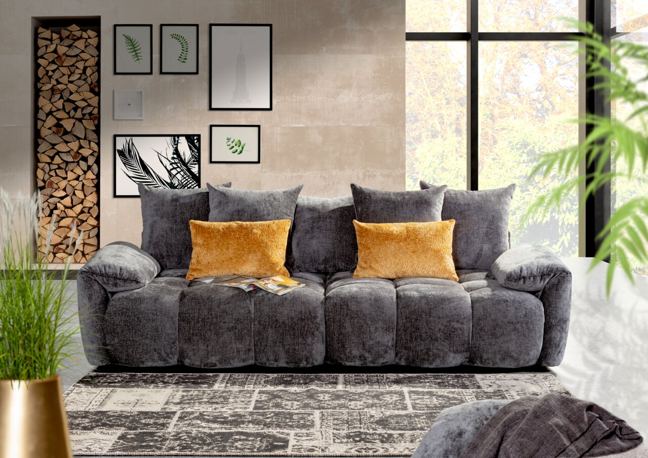 Graues Megasofa mit weichen Kissen und zwei gelben Zierkissen, frontal fotografiert in einem modernen Wohnzimmer mit großen Fenstern und Wanddekorationen.