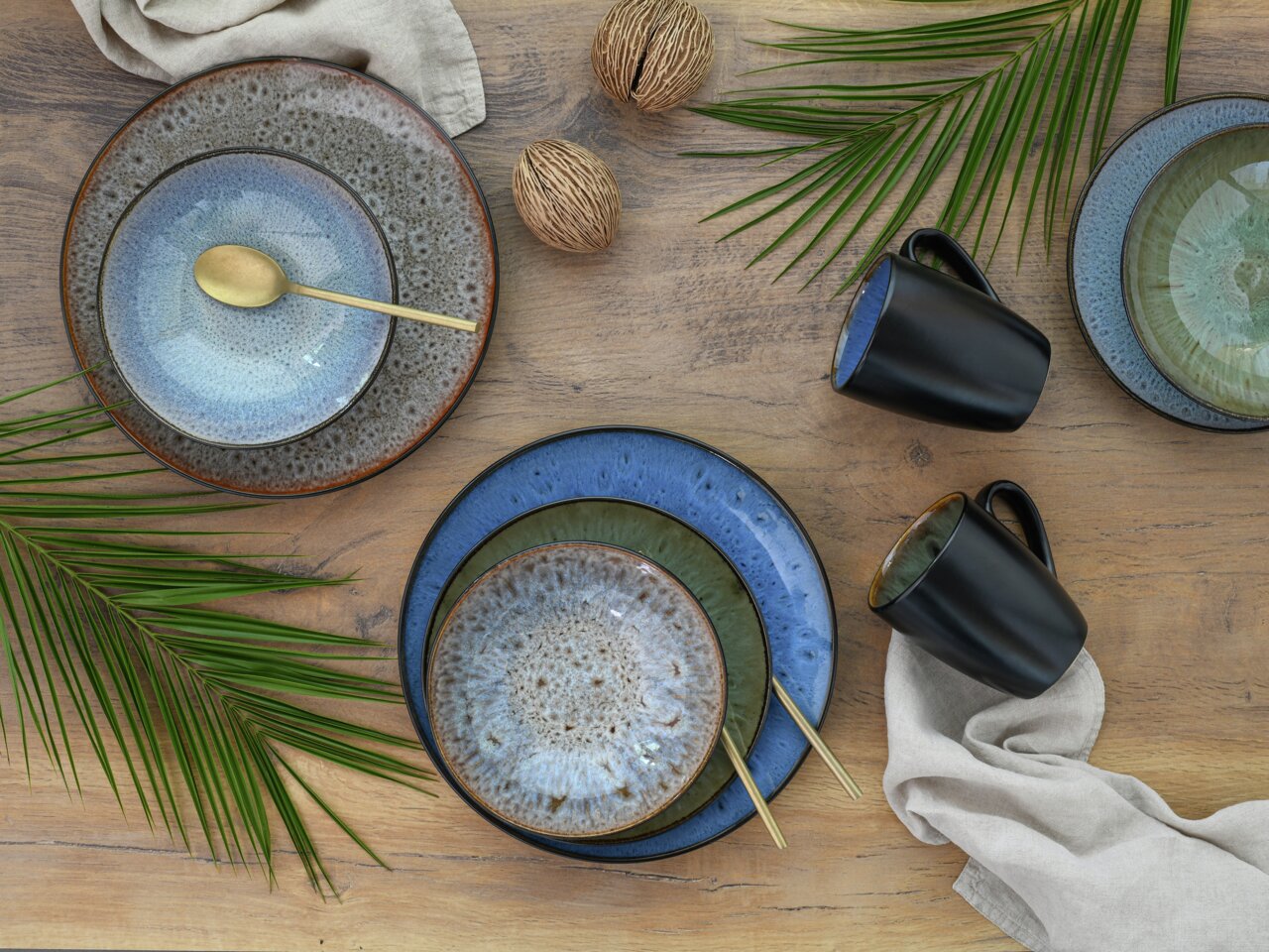 Vogelperspektive auf ein stilvolles Kombiservice Bali 16-tlg. mit Tellern in Blau- und Grüntönen, schwarzen Tassen und goldenen Löffeln, dekoriert mit Palmenblättern auf einem Holztisch.
