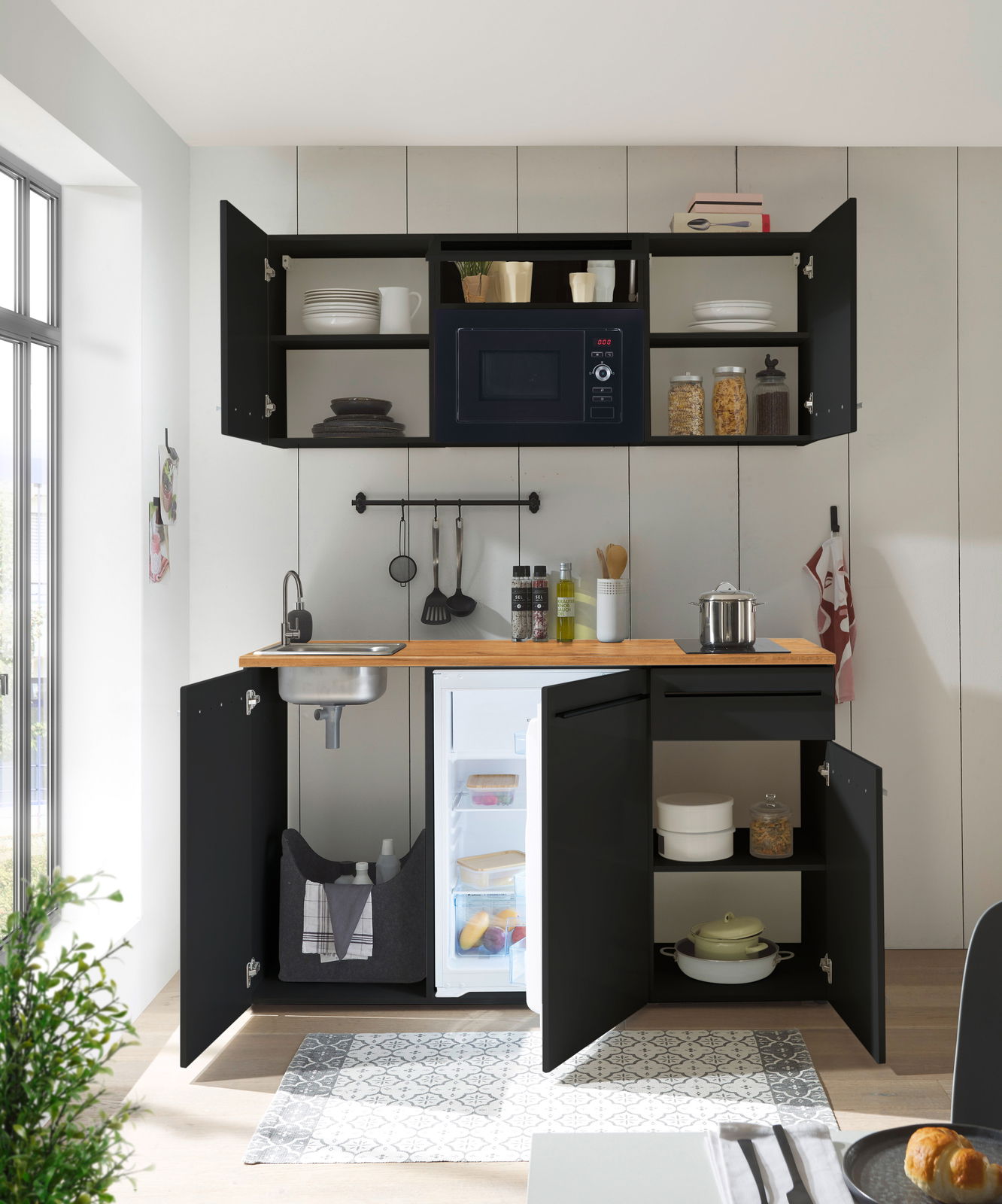 Moderne Single-Küche in schwarzem Design mit Holzarbeitsplatte, integriertem Kühlschrank, Spüle und Hängeschränken, aus der Perspektive von vorne betrachtet.