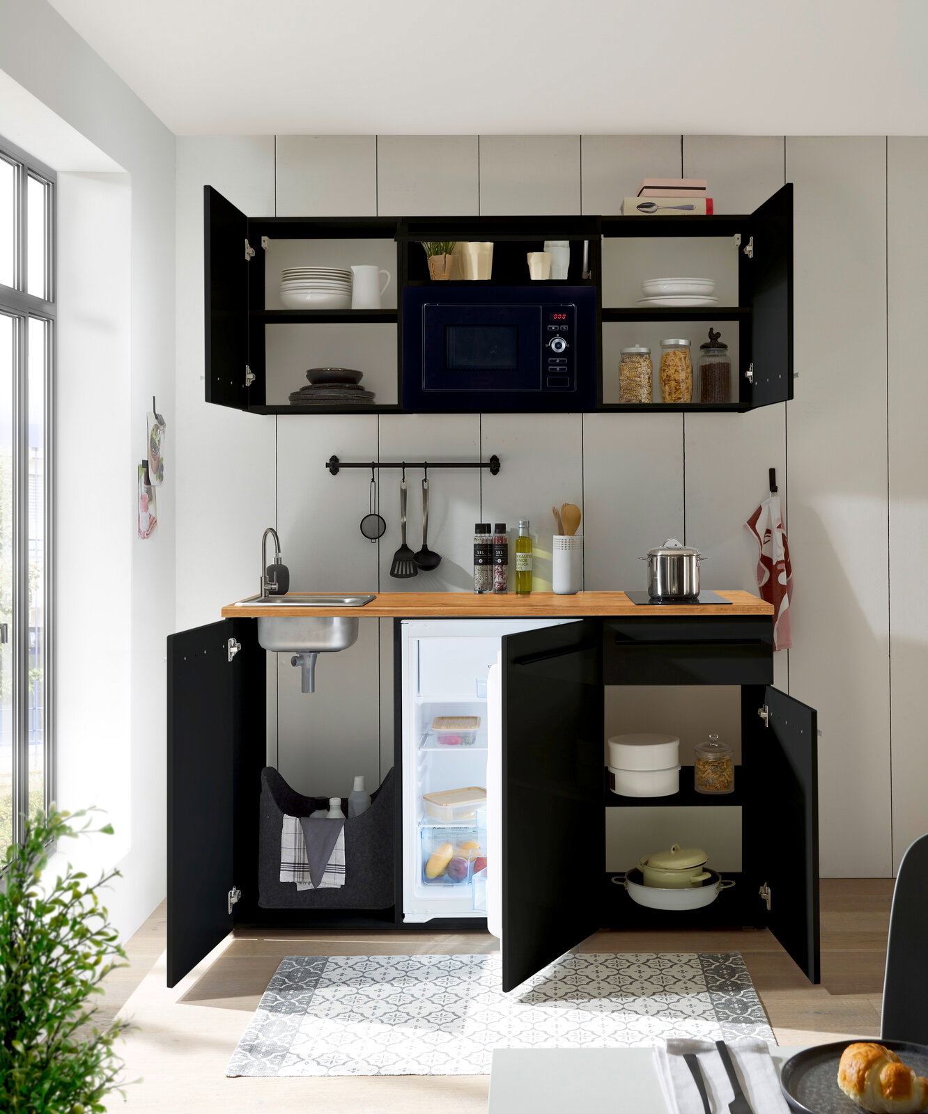 Moderne Single-Küche in schwarzem Design mit Holzarbeitsplatte, integriertem Kühlschrank, Spüle und Hängeschränken, aus der Perspektive von vorne betrachtet.