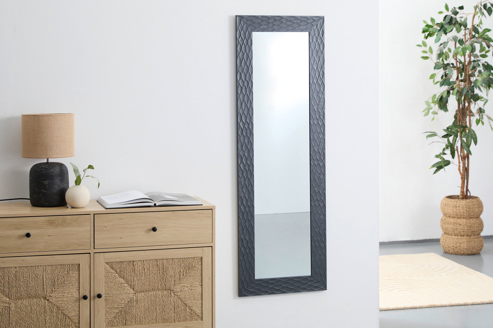 Rechteckiger Wandspiegel mit dunklem, strukturiertem Rahmen, aufgehängt in einem modernen Raum mit Kommode, Lampe und Pflanze, seitliche Perspektive.