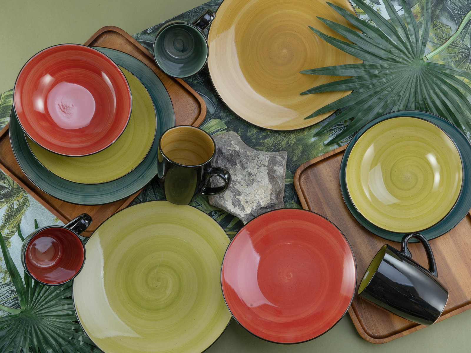 Vogelperspektive auf ein farbenfrohes 16-teiliges Kombiservice mit Tellern, Schalen und Tassen in Rot, Grün und Gelb auf einem tropischen Hintergrund.