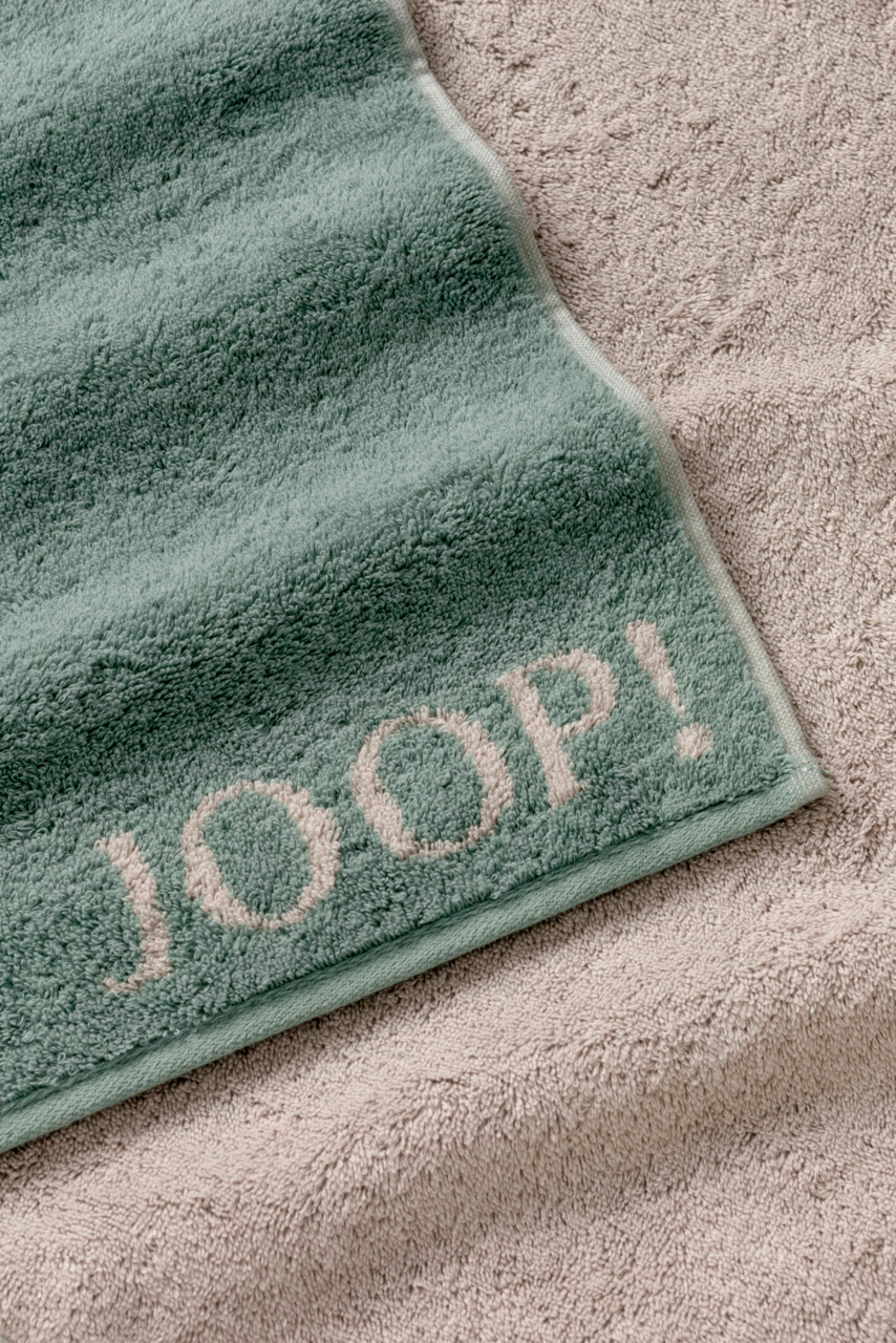 Grünes Handtuch mit dem Schriftzug 'JOOP!' in Nahaufnahme, auf einem beigen Handtuch liegend.