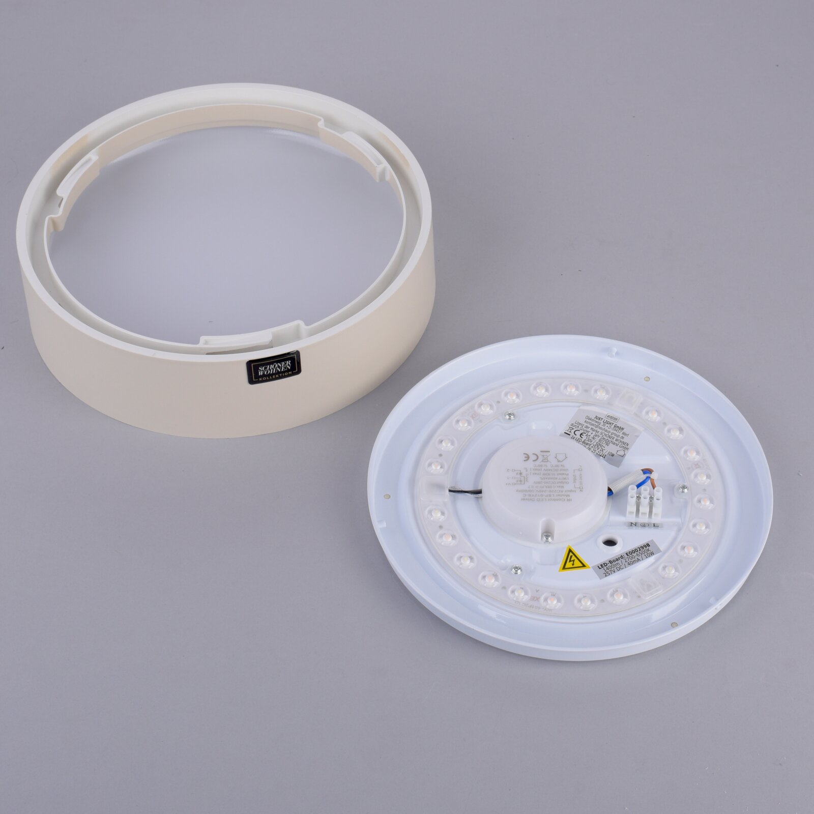 Weiße, runde LED-Deckenleuchte in Einzelteilen (Abdeckring und LED-Platine) auf grauem Hintergrund, schräg von oben fotografiert.