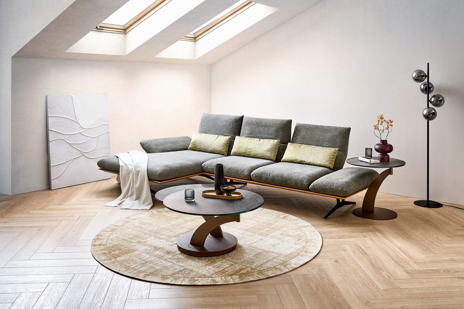 Modernes Wohnzimmer mit grauem Ecksofa und gelben Kissen, rundem Couchtisch und Beistelltisch auf hellem Teppich, aufgenommen in schräger Frontansicht aus leicht erhöhter Perspektive.