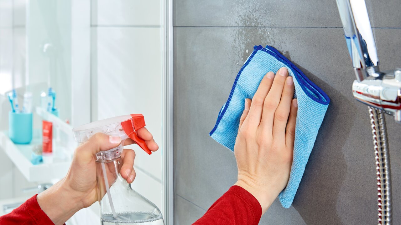 Eine Person reinigt eine Badezimmerfliese mit einem blauen Tuch und einem Sprühreiniger. Perspektive: Nahaufnahme der Hände bei der Reinigung.